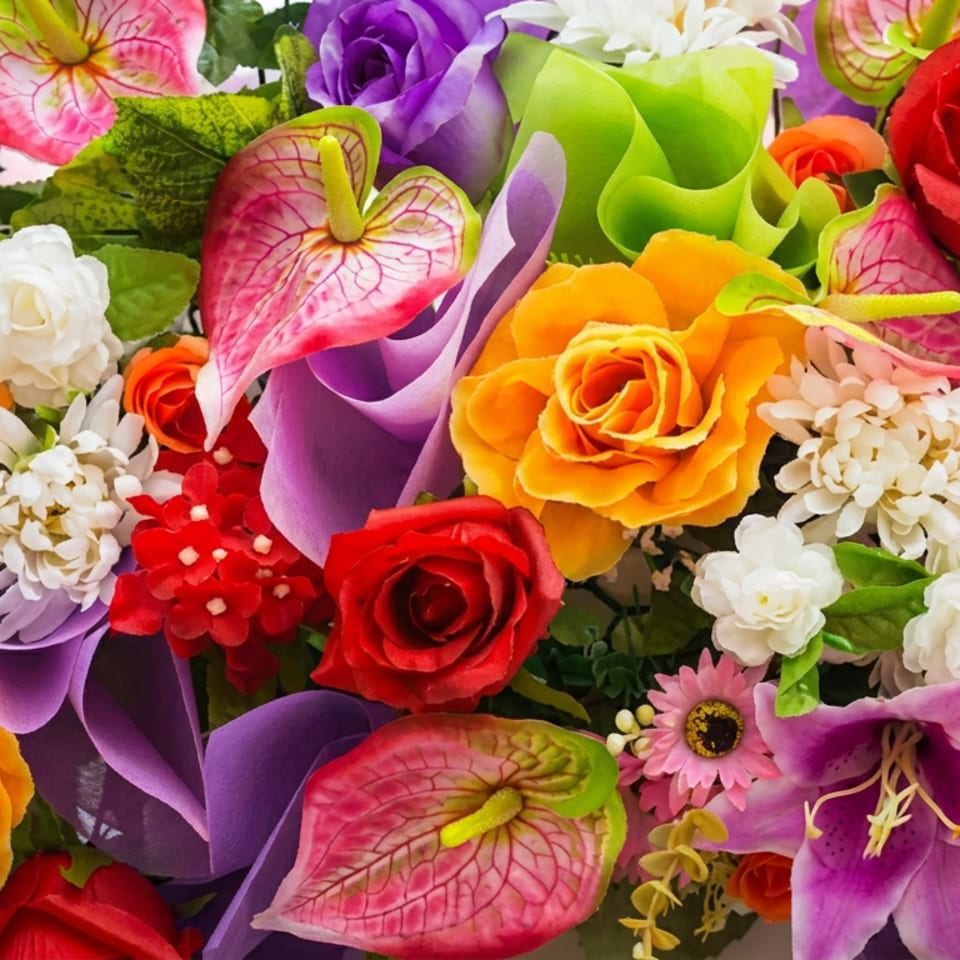 Colorful floral arrangement featuring red, orange, purple, pink, green, and white roses, lilies, anthuriums, and daisies.