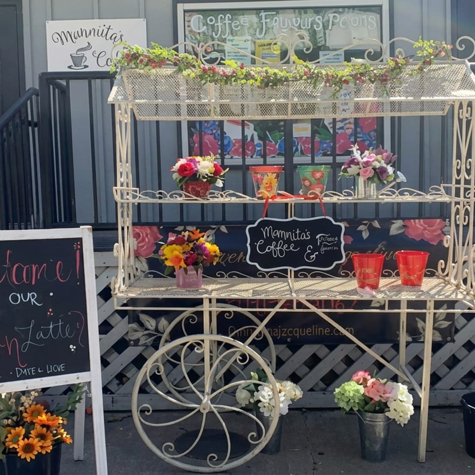 Outdoor coffee shop display with flowers, colorful cups, chalkboard advertising a latte, and café entrance behind it.